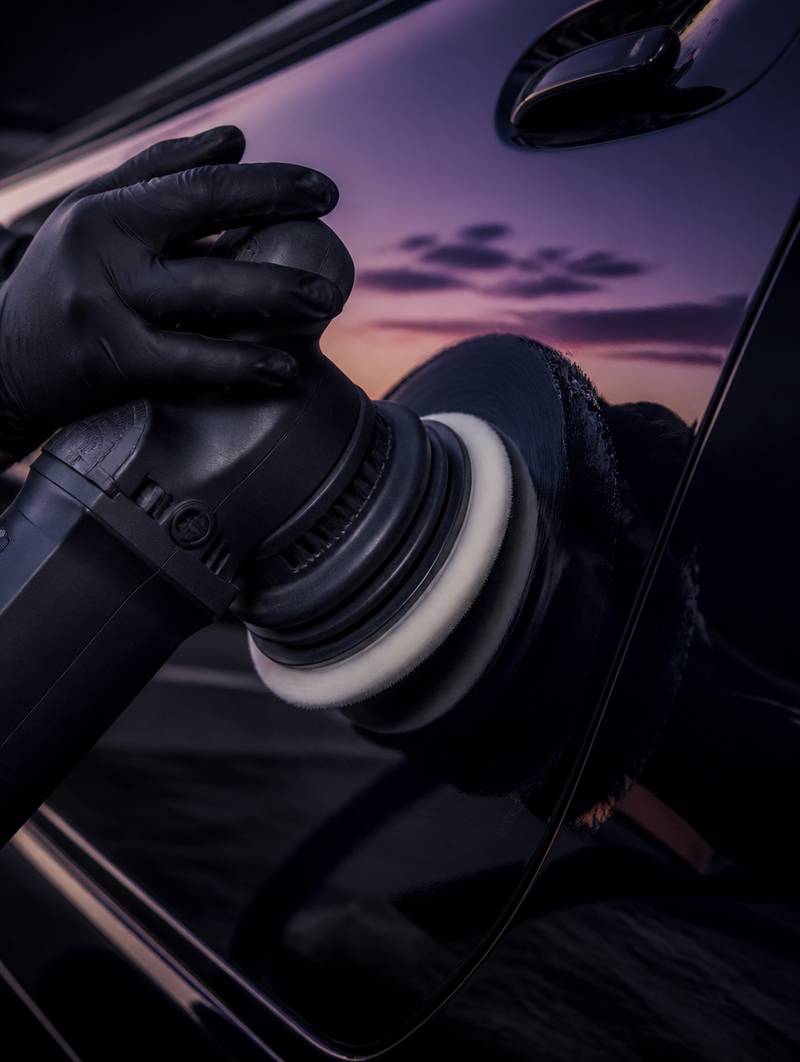 A detailer's hand guiding a dual-action polisher across glossy black paint